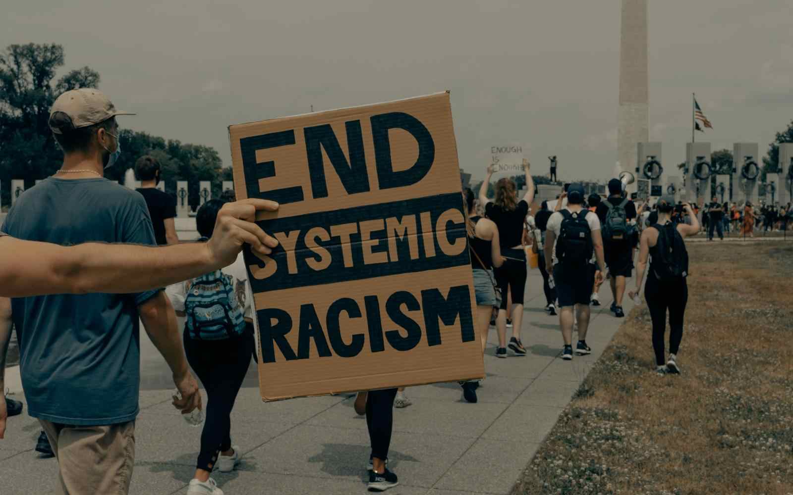 Karton z napisem "end systemic racism" (tłum. zakończmy systemowy rasizm) trzymana przez osobę protestującą. W tle inne osoby uczestniczące w proteście.