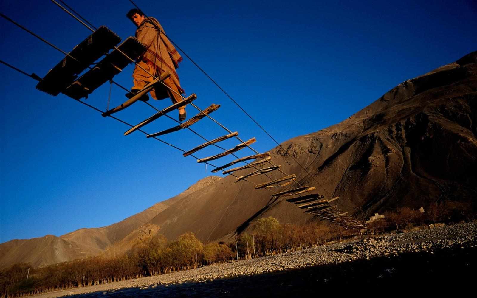 2020 Leading Social Entrepreneurs Front Cover. Depicting a man in the valleys of northeastern Afghanistan crossing a cable bridge with sparsely spaced wood planks above the Pandjchir River, connecting the village of Malespe to a refugee camp. Photographer and Ashoka Fellow, Reza Deghati, captured this shot in 2000.