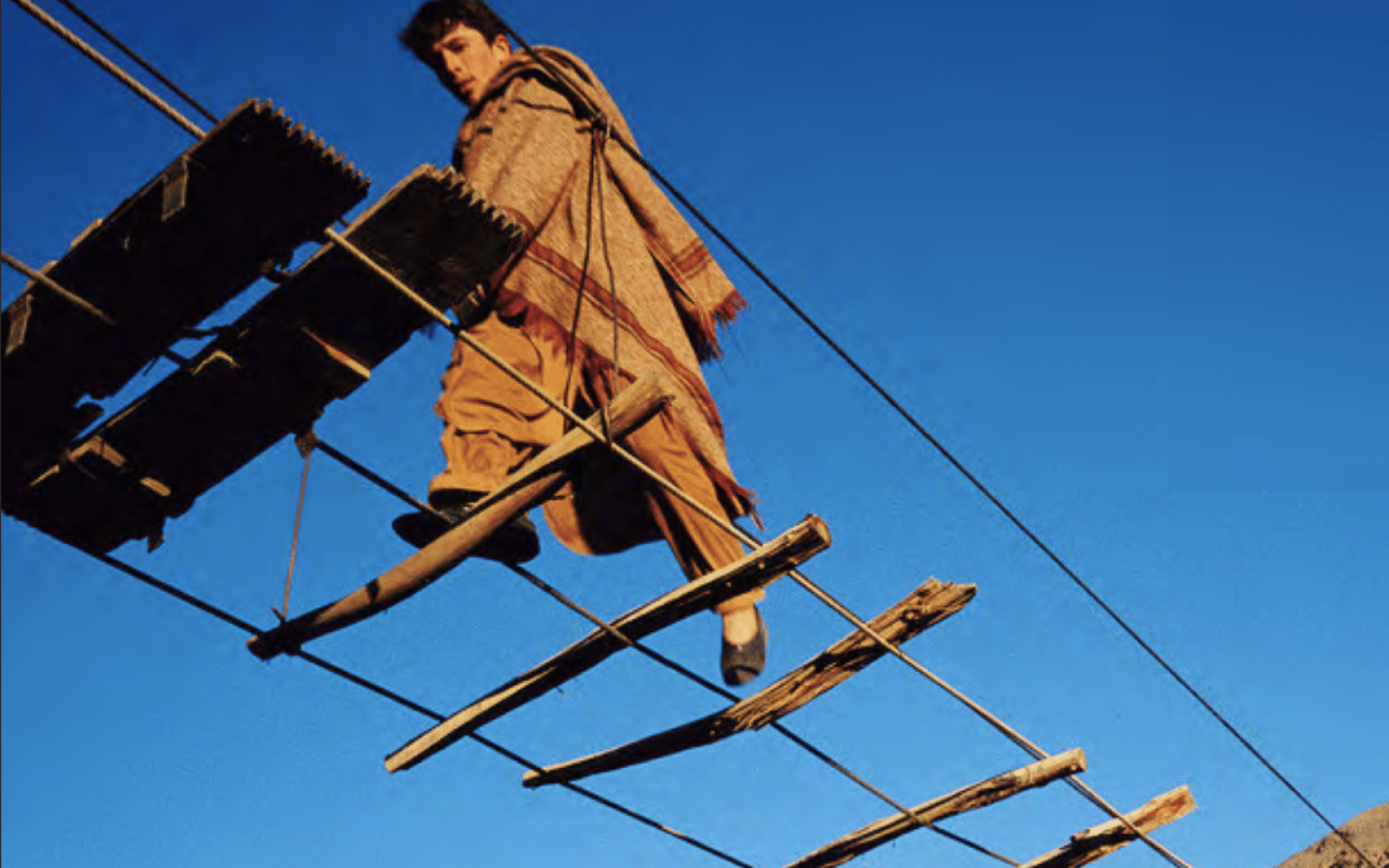Person in a cloak walking across a bridge that has minimal wooden boards on it