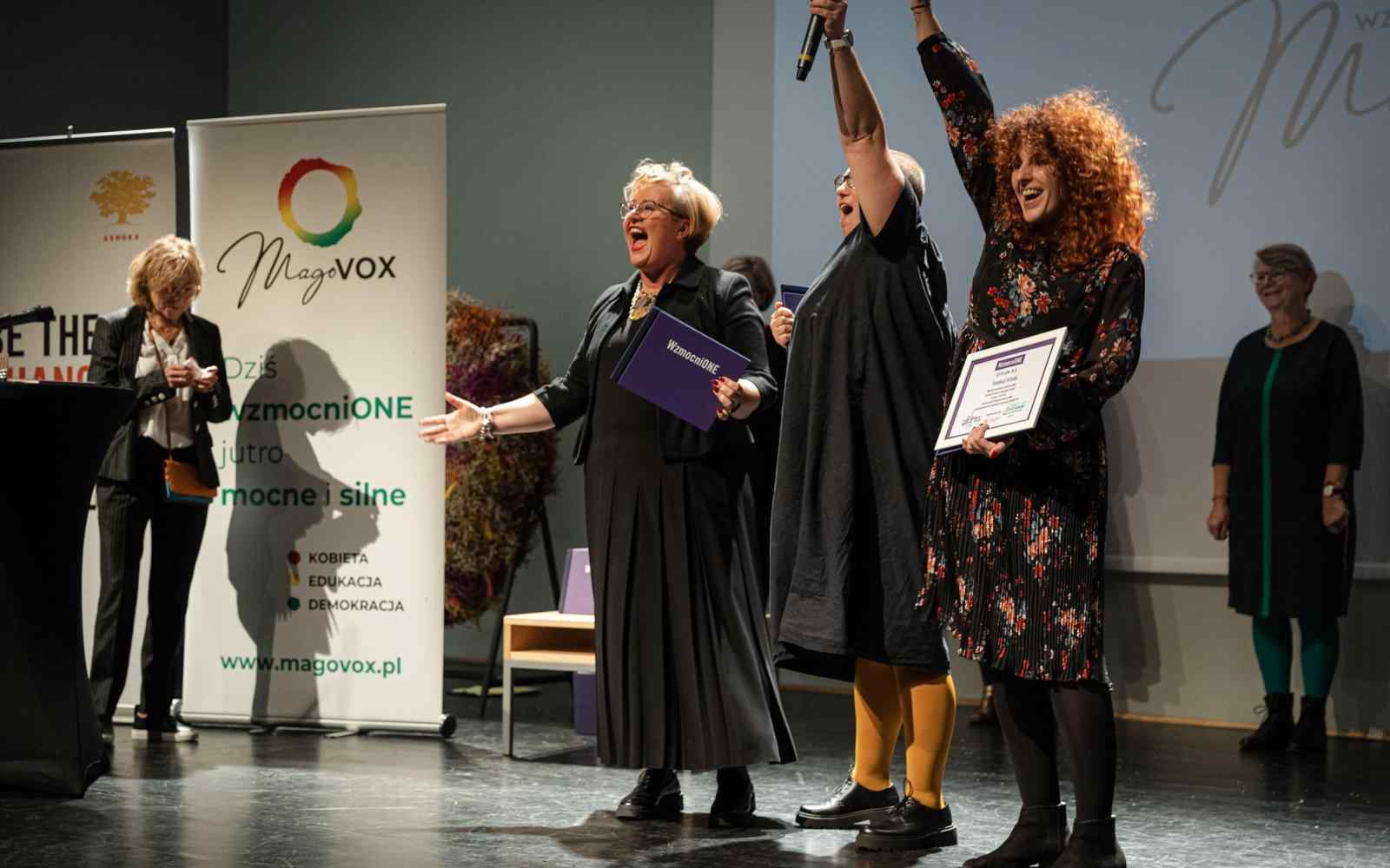 Zdjęcie przedstawia 3 członkinie Fundacji Oficyna-ToTutaj na scenie. Dwie podnoszą ręce w geście zwycięstwa, trzecia śmieje się z rozłożonymi rękami. W tle widać baner Ashoki i Magovoxu oraz uśmiechające się osoby z Jury programu.