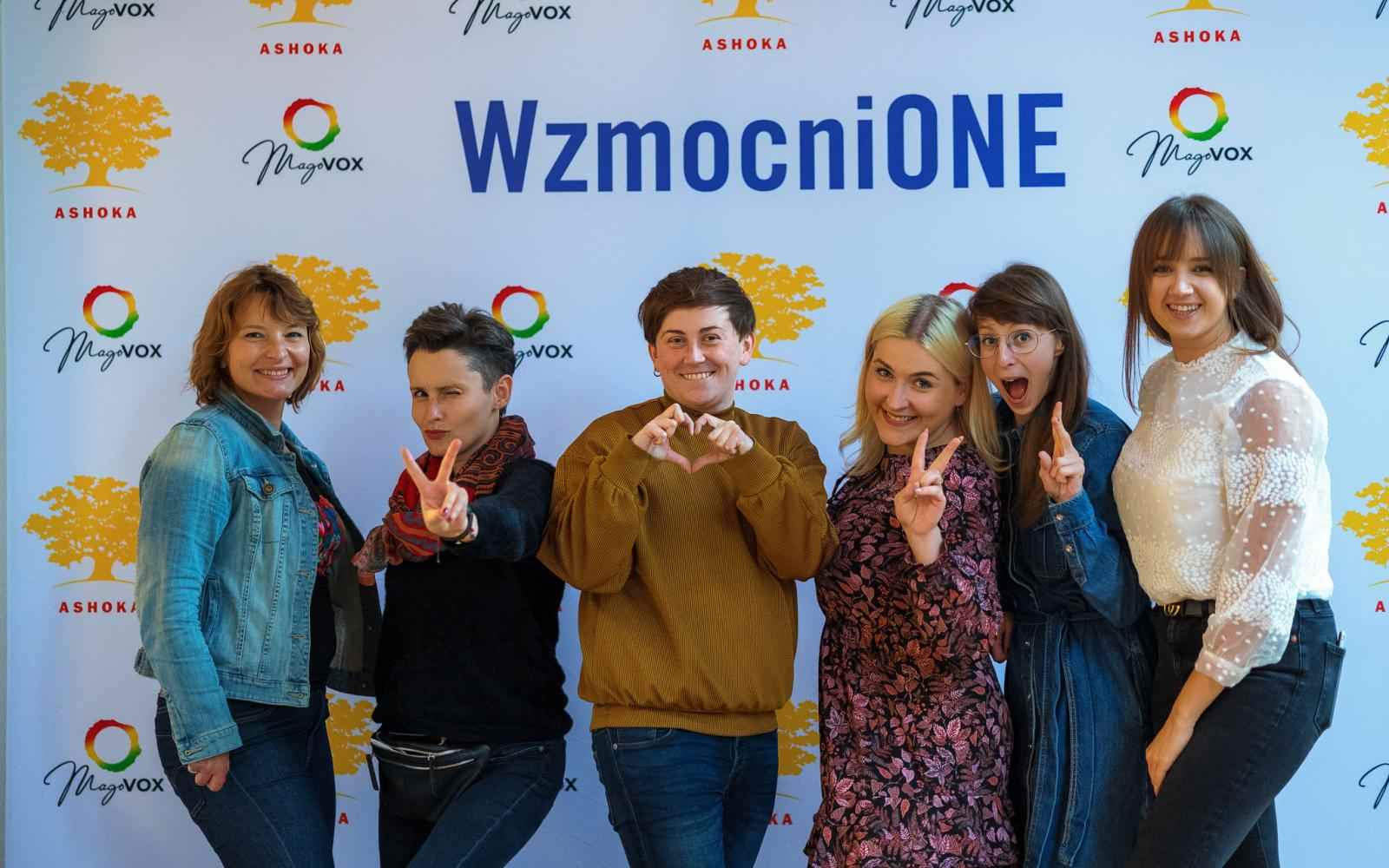 Na tle ścianki wzmocnionej z logo Wzmocnione, Ashoki oraz Magovox pozuje 6 osób w kolorowych ubraniach. Uśmiechają się, 3 pokazują znak peac/wolności palcami, jedna robi serduszko dłońmi.