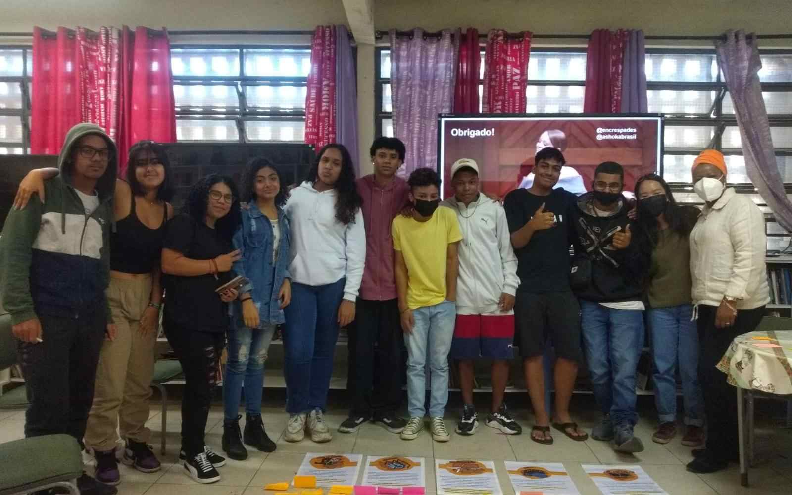 Jovens enfileirados posando para foto. No chão à frente do grupo, há folhas de papel e notas adesivas