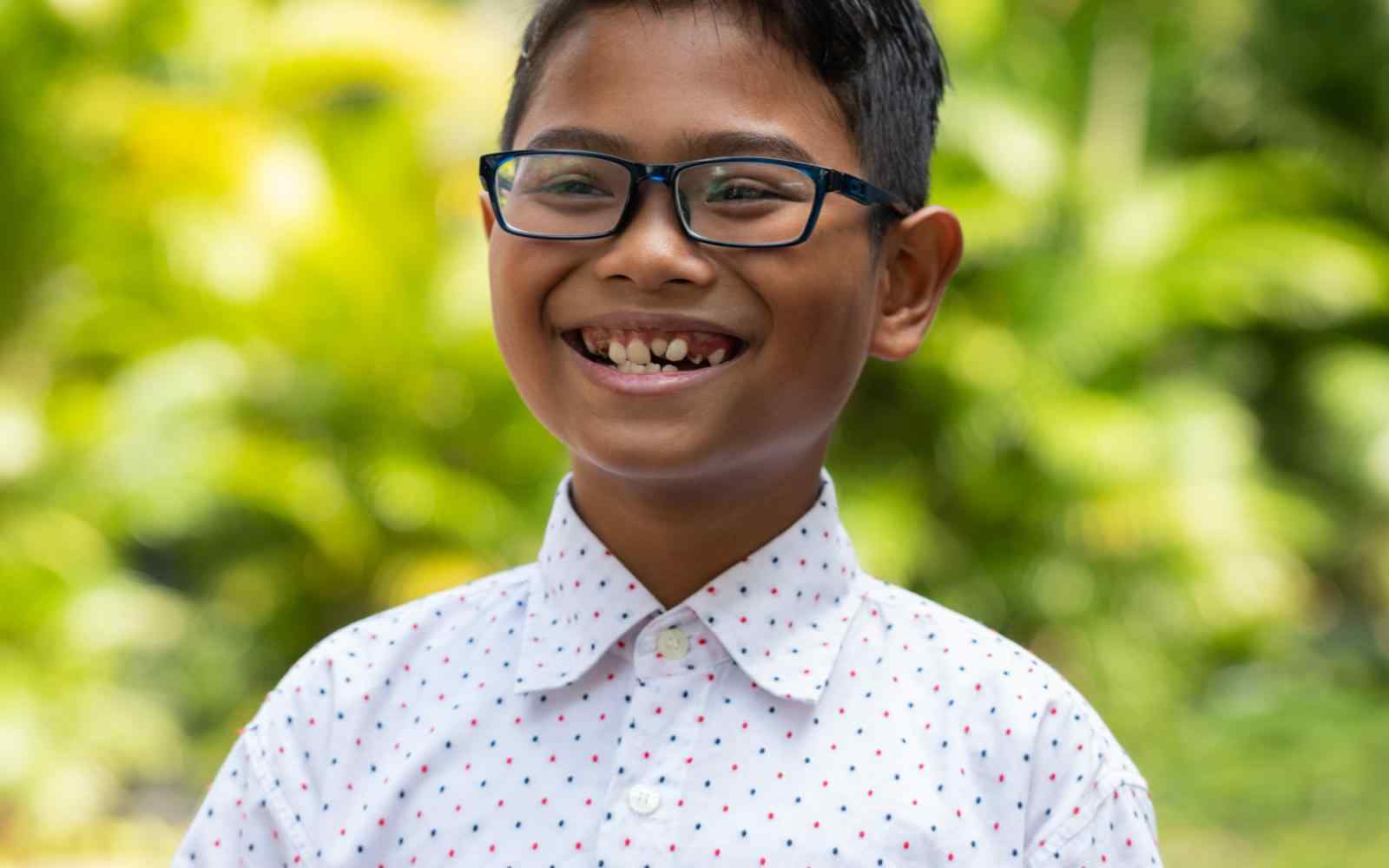 Headshot of AYC Bhre grinning cheerfully wearing a white polkadot shirt on a nature background.