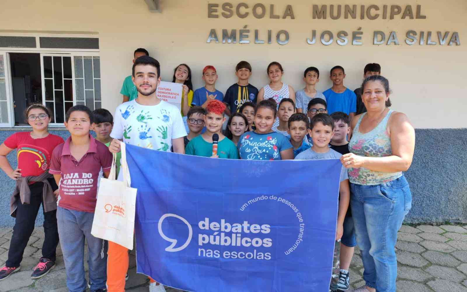 Grupo de crianças reunidas em frente à escola. Dois adultos estão junto com o grupo e seguram a bandeira dos Debates Públicos nas Escolas