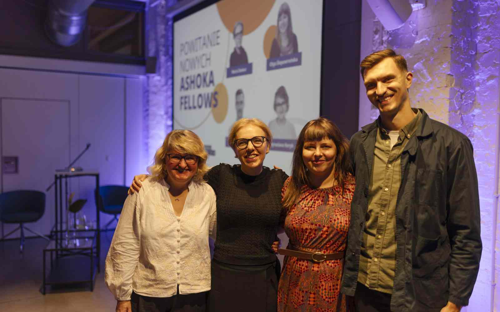 Zdjęcie przedstawia 4 Ashoka Fellows na spotkaniu witającym ich w społeczności Ashoki. Od lewej Myroslava Keryk, Maria Deskur, Olga Ślepowrońska i Maciej Otrębski. W tle widać filetowe światła i rzutnik.