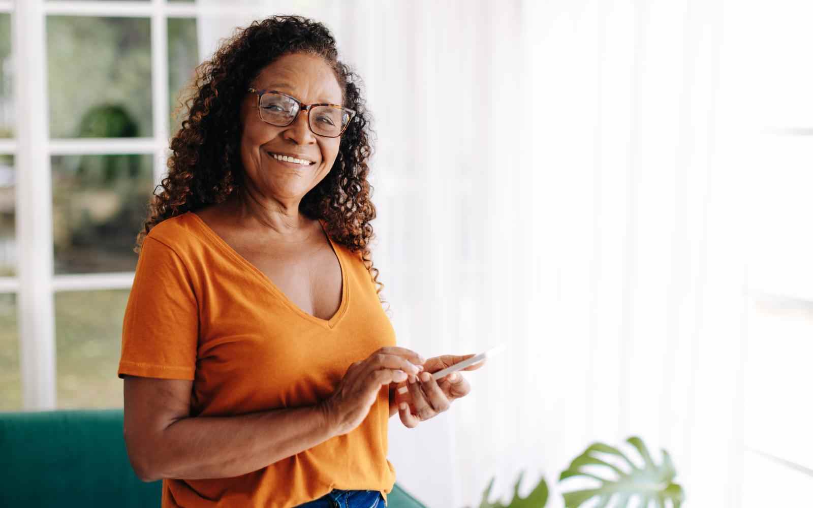 Mulher negra idosa de cabelo cacheado na altura dos ombros em uma sala de estar iluminada. Ela veste uma blusa laranja, segura um celular e sorri para a foto