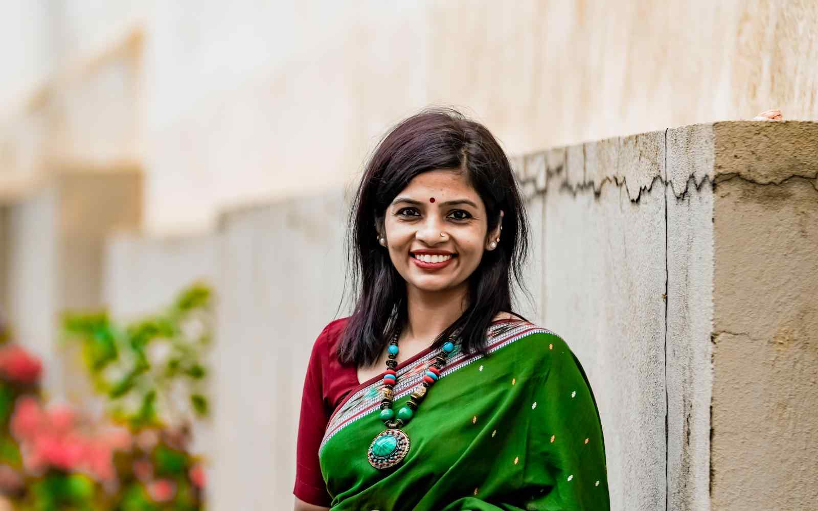 A headshot photo of Shruti Nair posing outside, in front of a stone wall with flowers