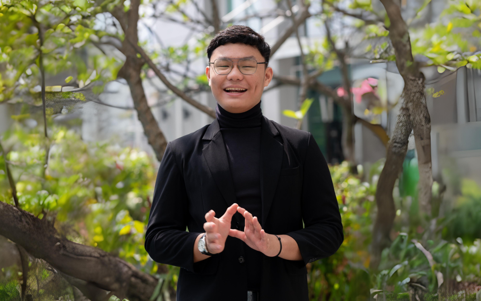 Headshot of AYC Atha smiling at the camera wearing glasses and a black suit on a nature background.