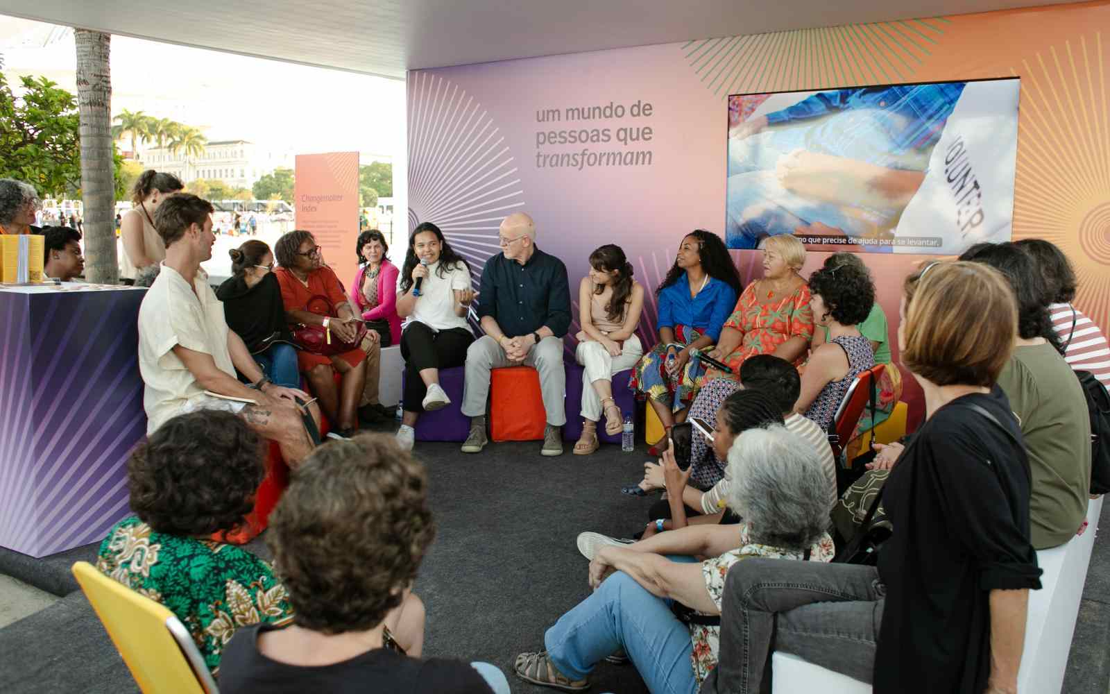 Grupo de pessoas reunida em um estande ao ar livre. Elas estão sentadas em roda, em pufes, bancos e cadeiras de praia. Ao fundo, há um painel colorido onde se lê "um mundo de pessoas que transformam" com uma televisão ao lado