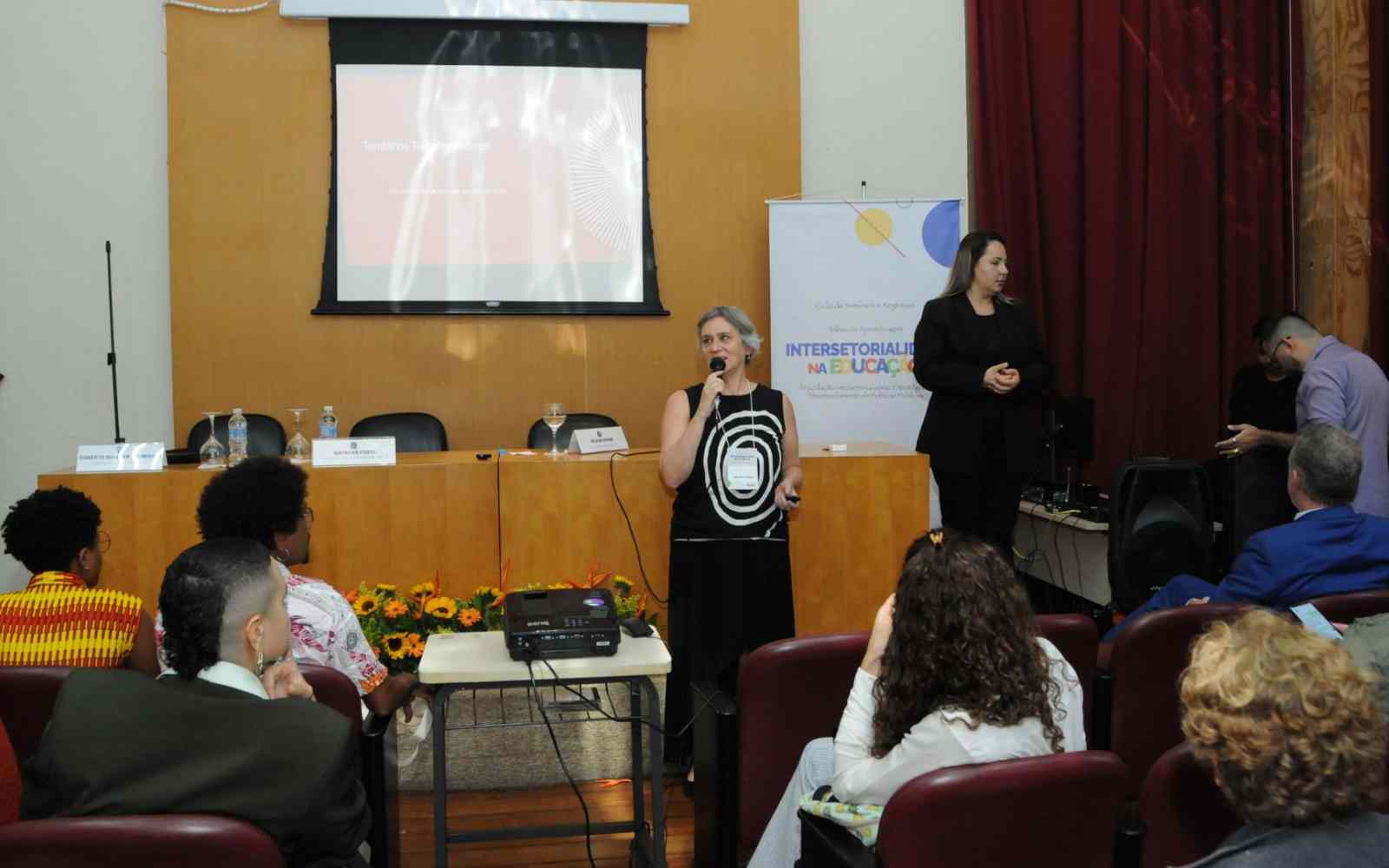 Vê-se Helena Singer (mulher branca de cabelos médios, lisos e grisalhos) segurando um microfone em frente a uma mesa em cima de um palco com diversas pessoas sentadas em cadeiras de frente para ela