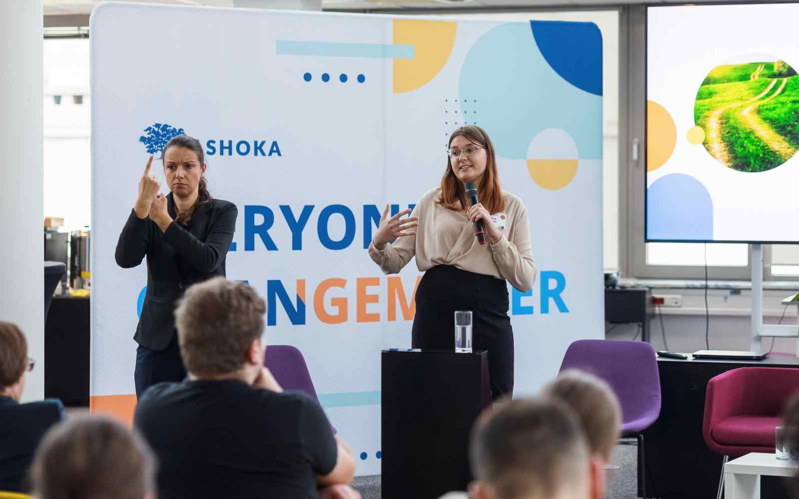 A woman speaking on stage, next to her another woman translating to sign language