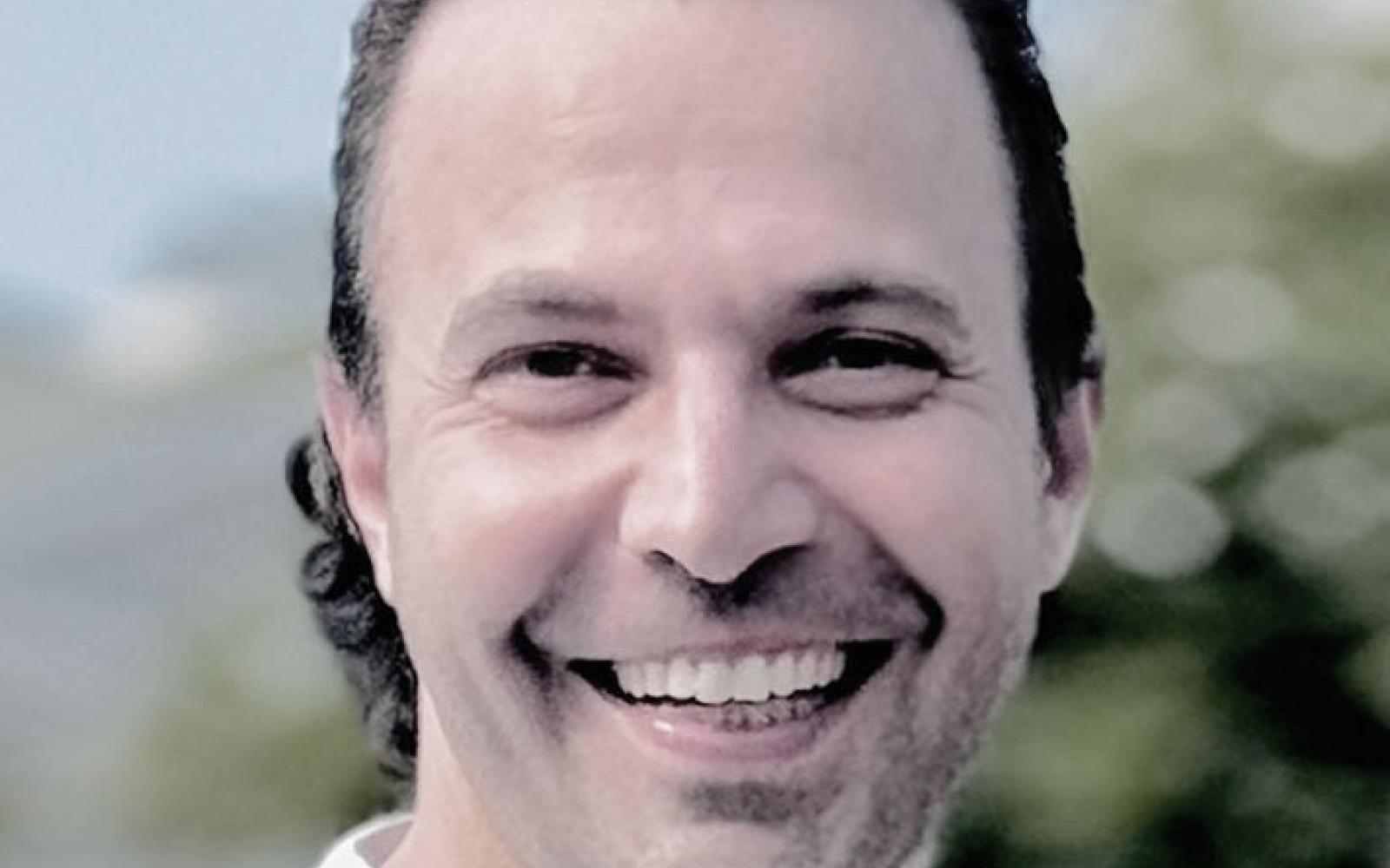 Profile photo of Ashoka Framework Change Staff Edward Edilbi. Photo of person with long slick backed hair smiling at the camera; background is of blurred out trees