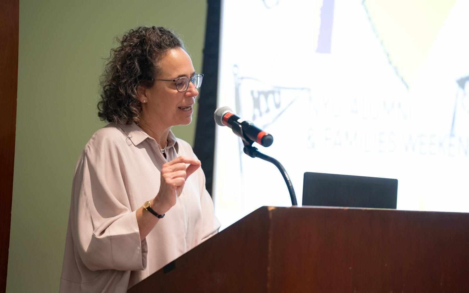 Photo of Diana Wells accepting GSAS Distinguished Alumni Achievement Award. Person with dark curly shoulder length hair and glasses speaking into a microphone attached to a dark brown podium. Dressed in light pink blouse; background is the front of a what appears to be a classroom or auditorium