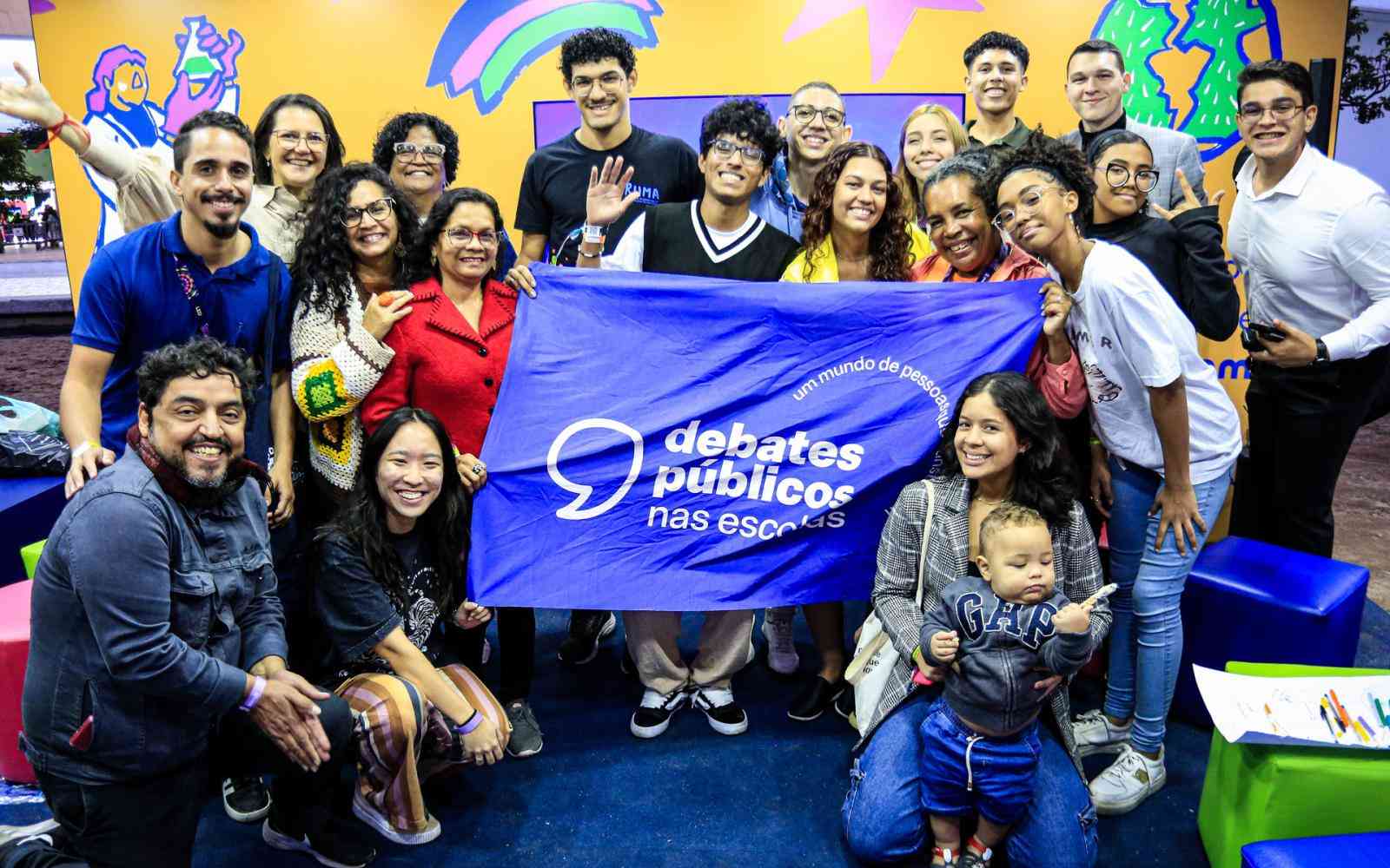 Grupo de pessoas de diferentes idades reunidas em um stand ao ar livre. Em destaque, elas seguram a bandeira dos Debates Públicos nas Escolas. Ao fundo, há um painel laranja com ilustrações coloridas.