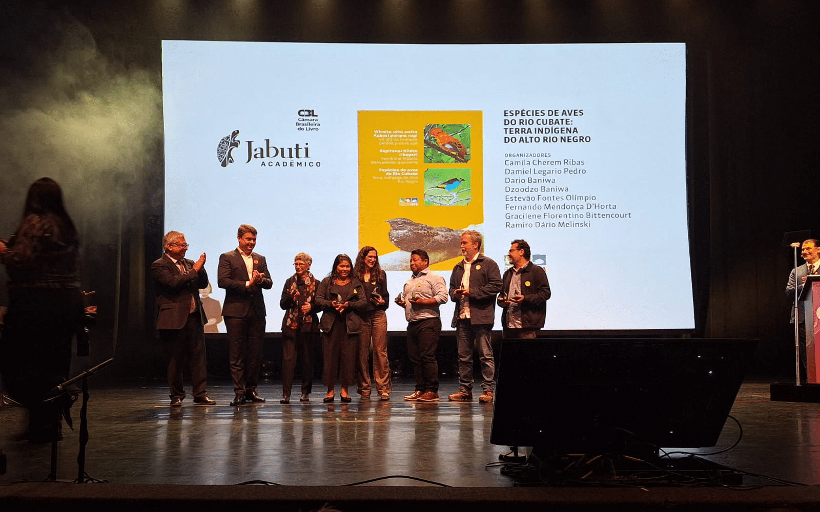 Pessoas em pé sobre um palco, algumas aplaudindo, outras sorrindo e segurando troféus. Ao fundo, um telão projeta a capa do livro “Espécies de Aves do Rio Cubaté: Terra Indígena do Alto Rio Negro”, com ilustrações de aves coloridas. No canto esquerdo da projeção, aparecem os logos da Câmara Brasileira do Livro e do Prêmio Jabuti Acadêmico.
