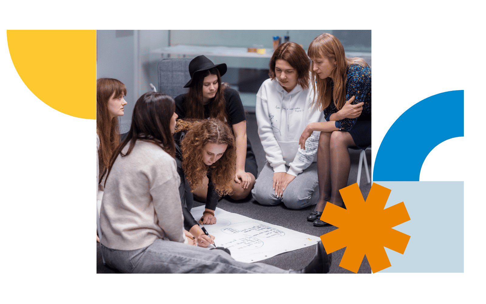 A group of six people is sitting on the floor in a modern indoor setting, gathered closely around a large sheet of white paper placed in the center. The paper appears to have diagrams or handwritten notes on it. The individuals are dressed casually; one person is wearing a black hat, and another is in a white sweatshirt. They are leaning forward, actively engaged with the content on the paper, suggesting a collaborative activity or brainstorming session.