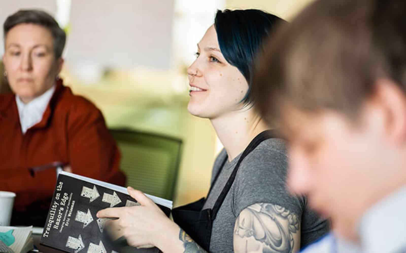 Three people sitting in a classroom setting; one person with lighter skin and dark blue hair smiling to the left of the photo.