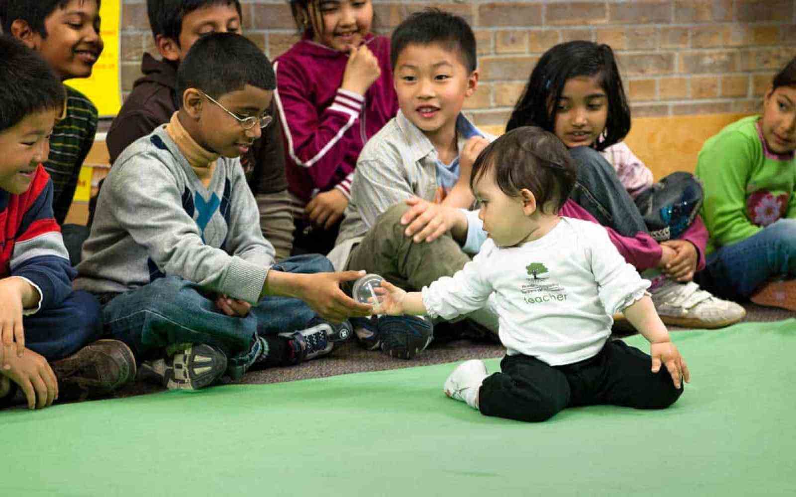 Baby handing an elementary school aged child an object while other students look on; located in a classroom
