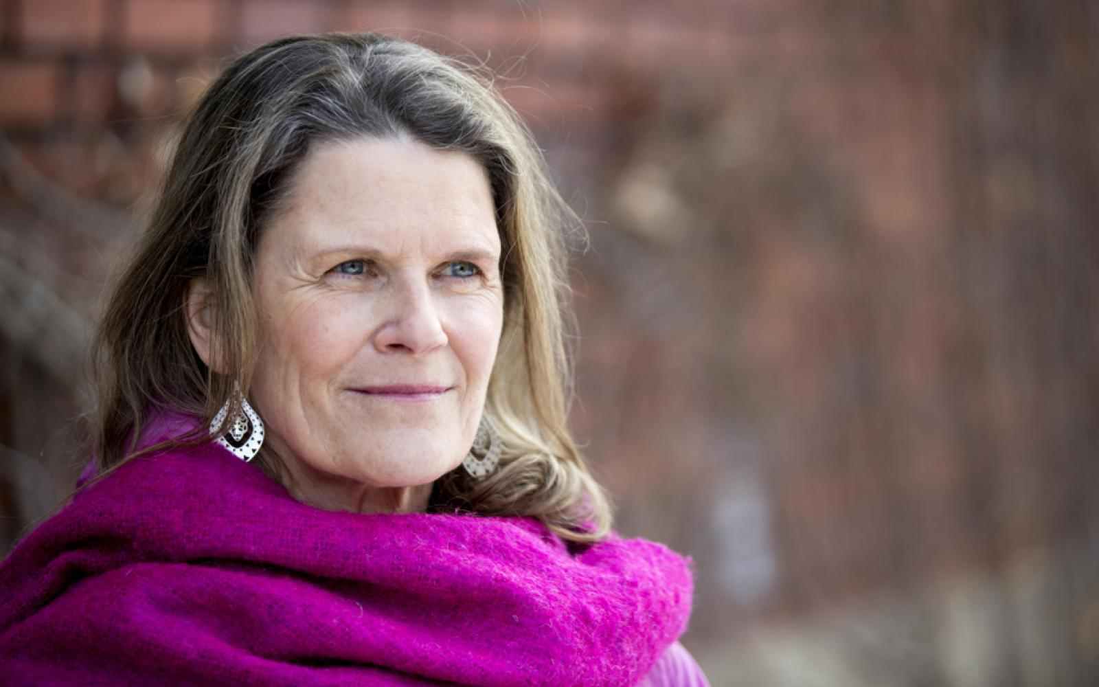Photo of Canada Fellow Rachel Kiddell-Monroe. Photo of a person with longer brown hair and lighter skin smiling to the right of the camera. Dressed in a pink scarf; background of a brick wall.