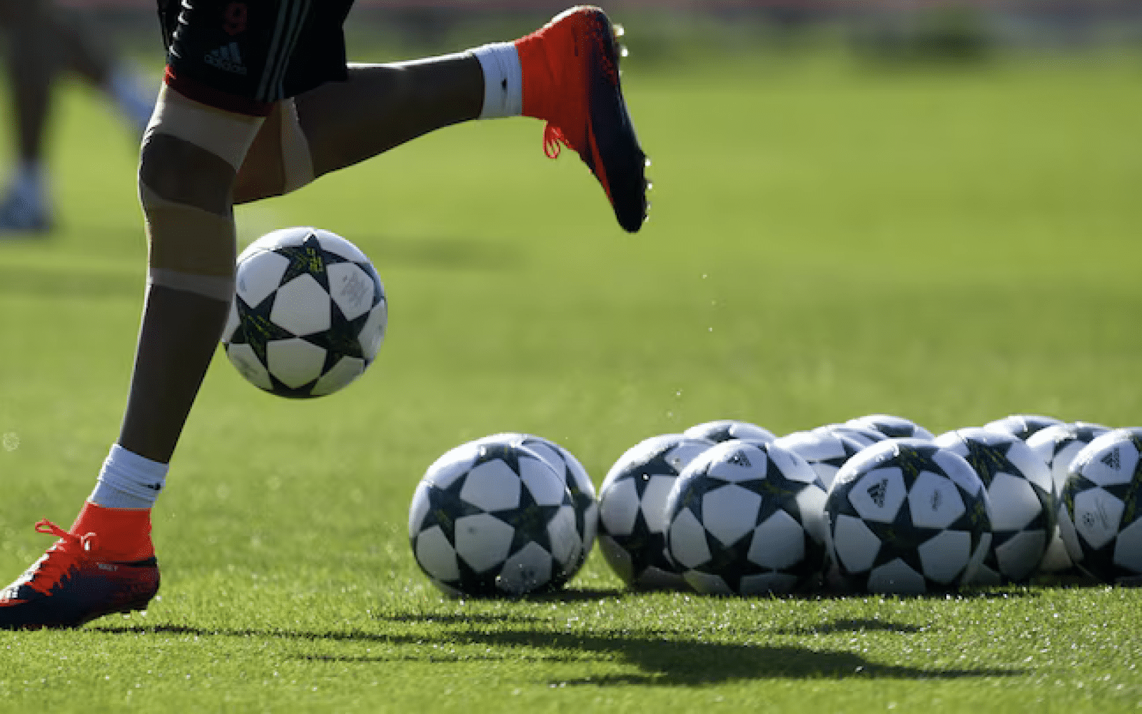 Photo of bottom half of soccer player's body  running on a field; next to him are a number of soccer balls