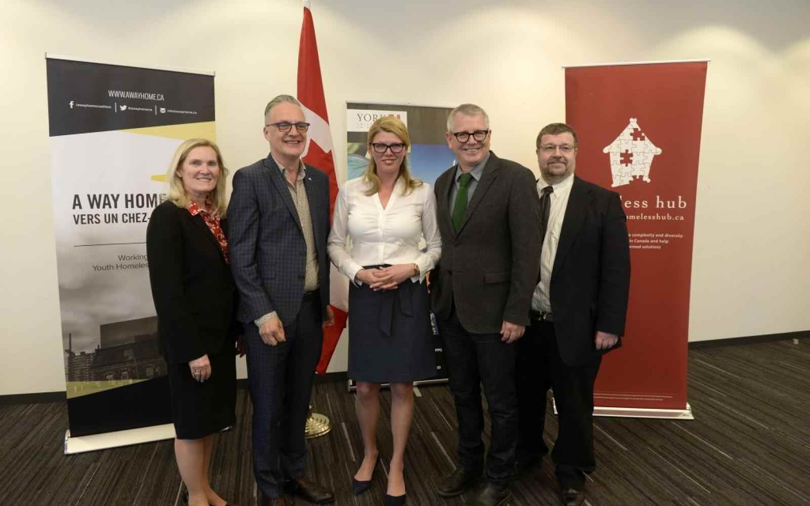 Five people dressed in suits smiling at the camera; backdrop of a few posters relating to the Federal funding for youth homelessness in Canada
