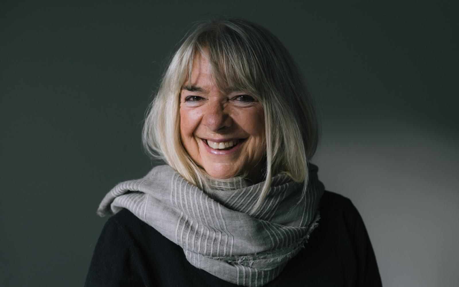 Person with shoulder length white hair smiling to the left of the camera. Background a white wall