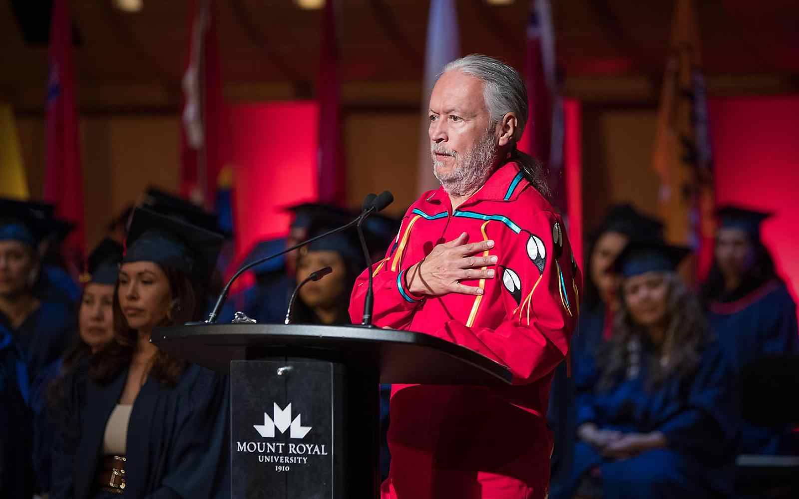 Person with grey hair and beard dressed in a red formal long shirt with indigenous art on it holding hand over heart and speaking to a graduation on a stage