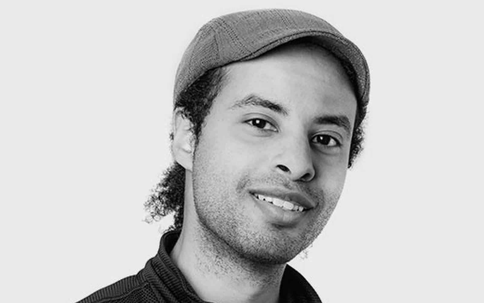 Person with fedora and short dark hair smiling at the camera. Photo in black and white
