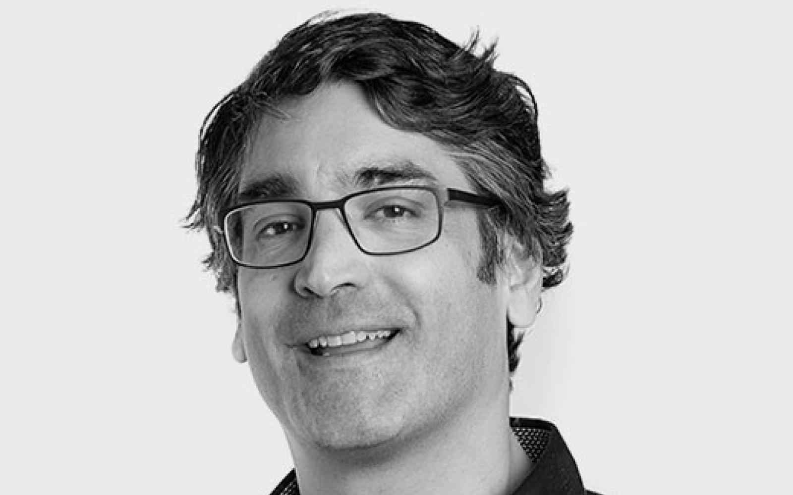 Headshot of Anil Patel, Ashoka Fellow in Canada. Black and white photo of a person with wavy hair and glasses smiling at the camera. Background is white wall