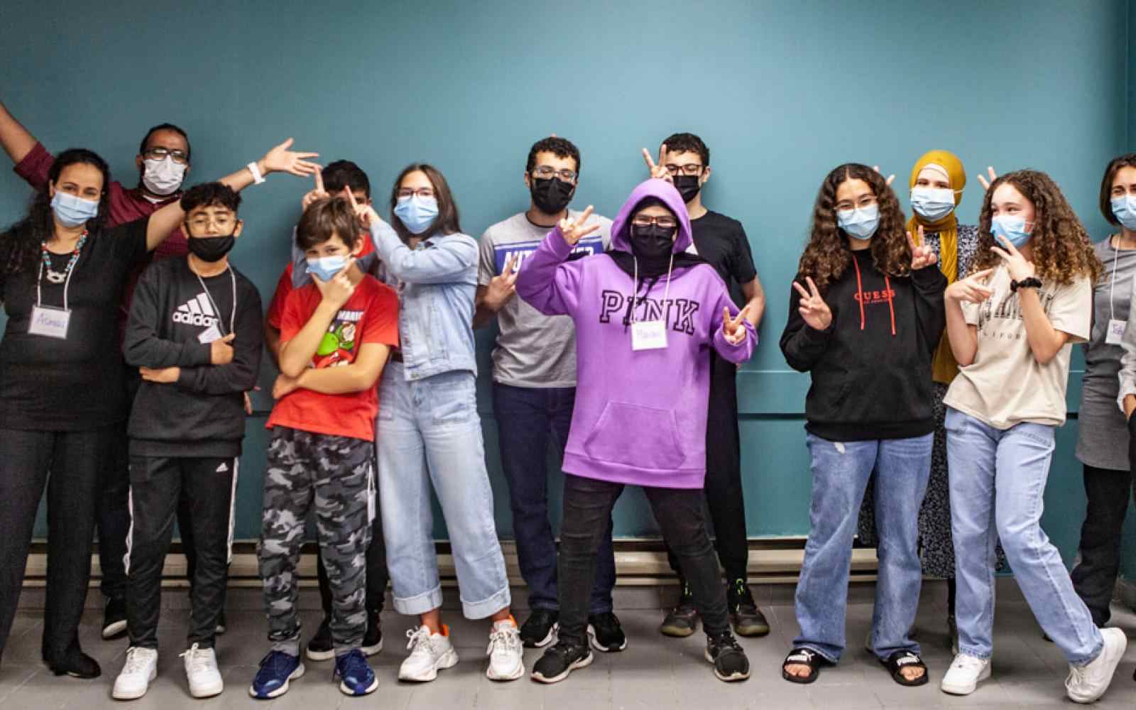 Group of kids with masks on smiling at the camera in a hallway; wallpaper is bright green-blue color