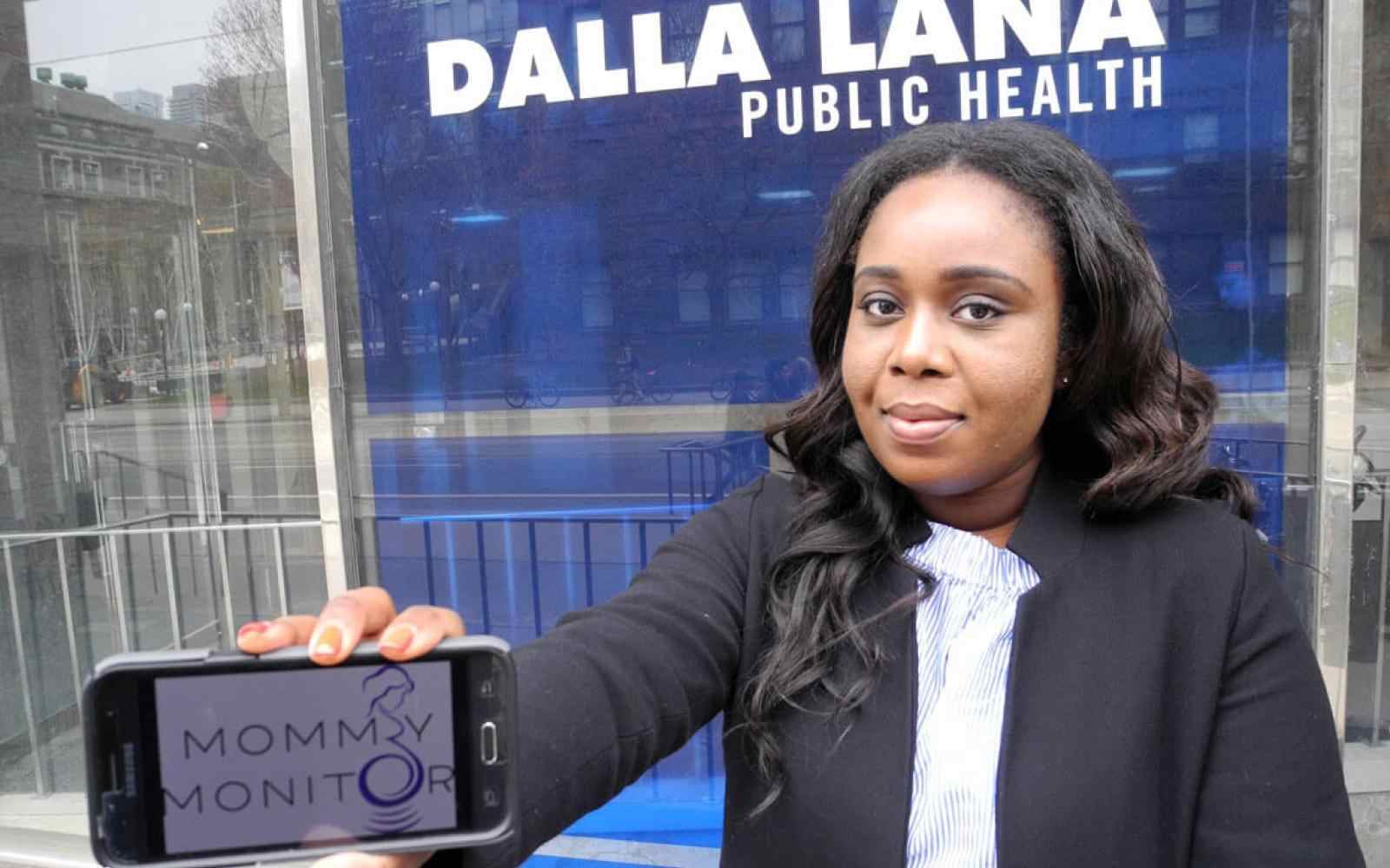 Person with short dark hair and brown skin smiling at the camera, dressed in a business suit. Showing a cell phone horizontally to the camera that has a logo for "Mommy Monitor" on it. Background is Dalla Lana Public Health school at U Toronto