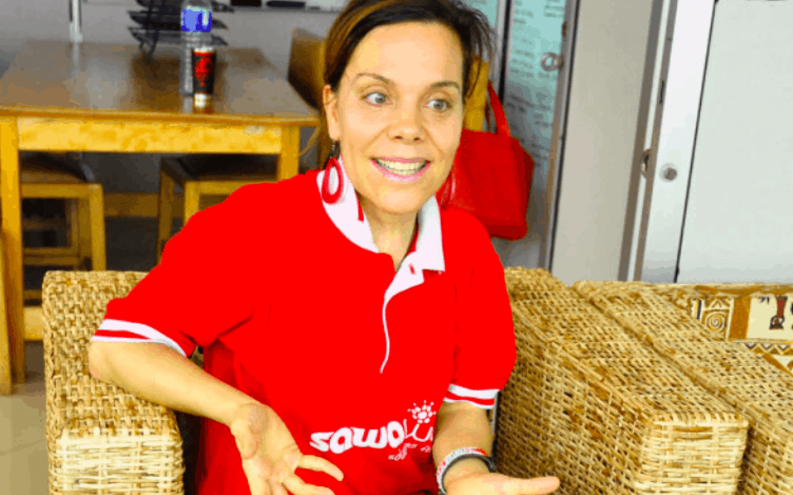Photo of Canada Fellow Daphne Nederhorst; person with short dark hair seated in a wicker chair explaining something to the camera. Has a red Sawa World t shirt on