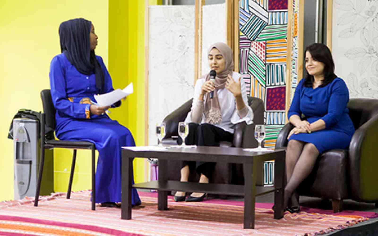 Two young Muslim women speaking on a stage as part of a panel. Background a lifted stage and a few banners