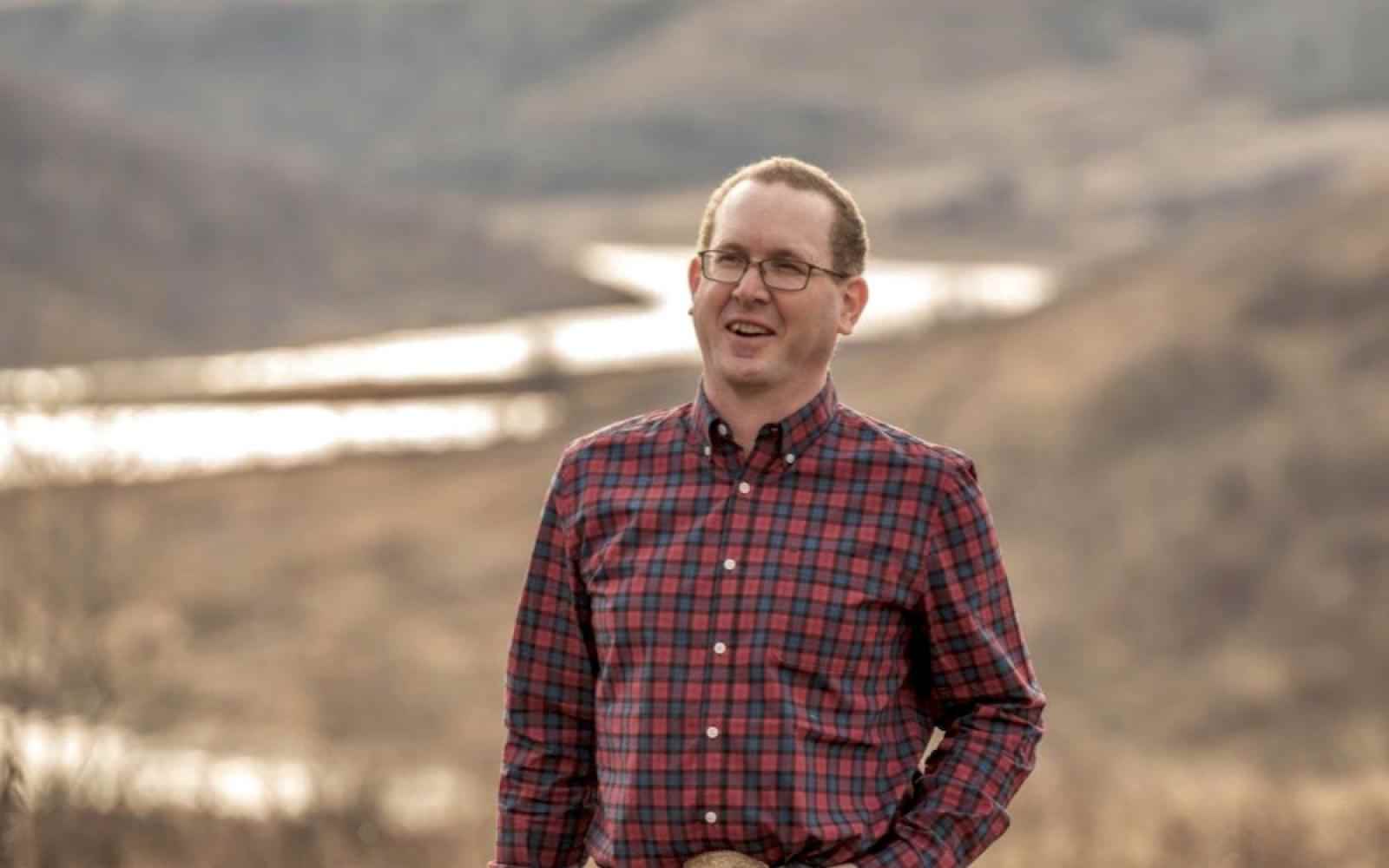 Photo of Sean Mcgrath, rancher in Canada with ALUS. Person with short dark hair, glasses, and a flannel shirt smiling at the camera; background is a river between hills