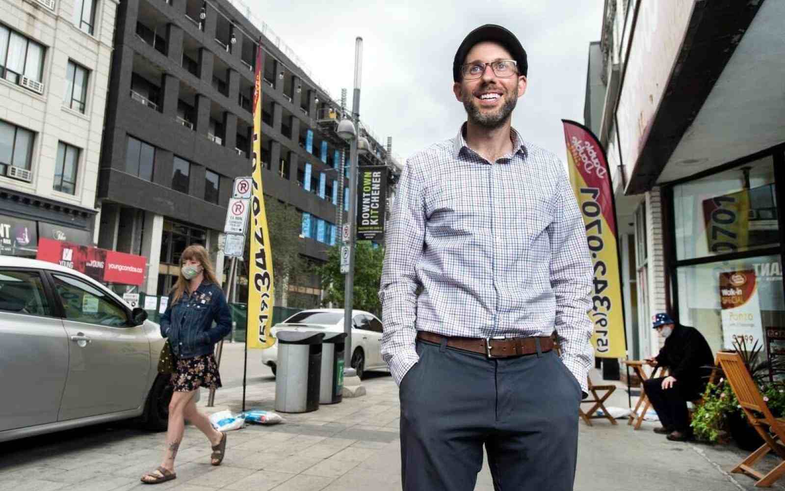 Person with a news cap on dressed in formal dresswear smiling at the camera backdrop of a sidewalk leading down a street behind the person