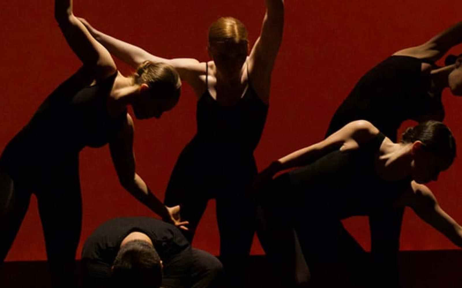 Picture of 5 dancers dressed in black tight dancing attire in a low light with red background arranged closely to each other mid choregraphy