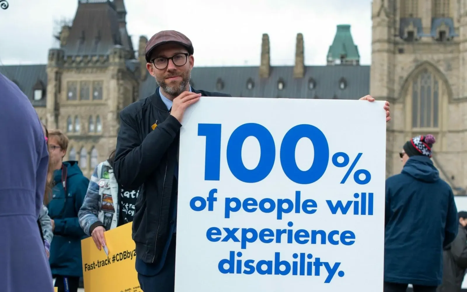 Person with a sign that says 100% of people will experience disability, background an old building below a skyline