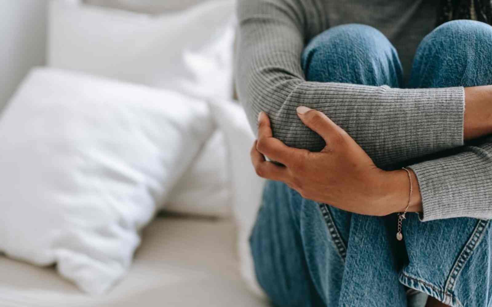 Person with femme attributes holding her knees on a bed; face off camera
