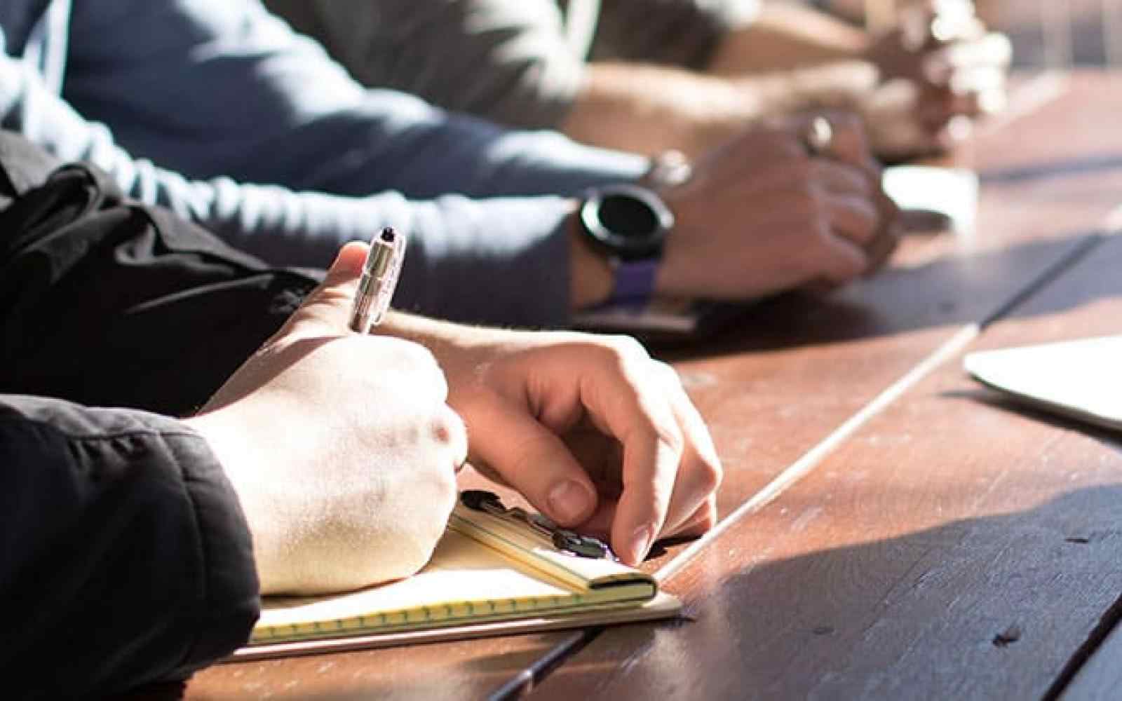 Photo of people's hands writing at a long wooden desk