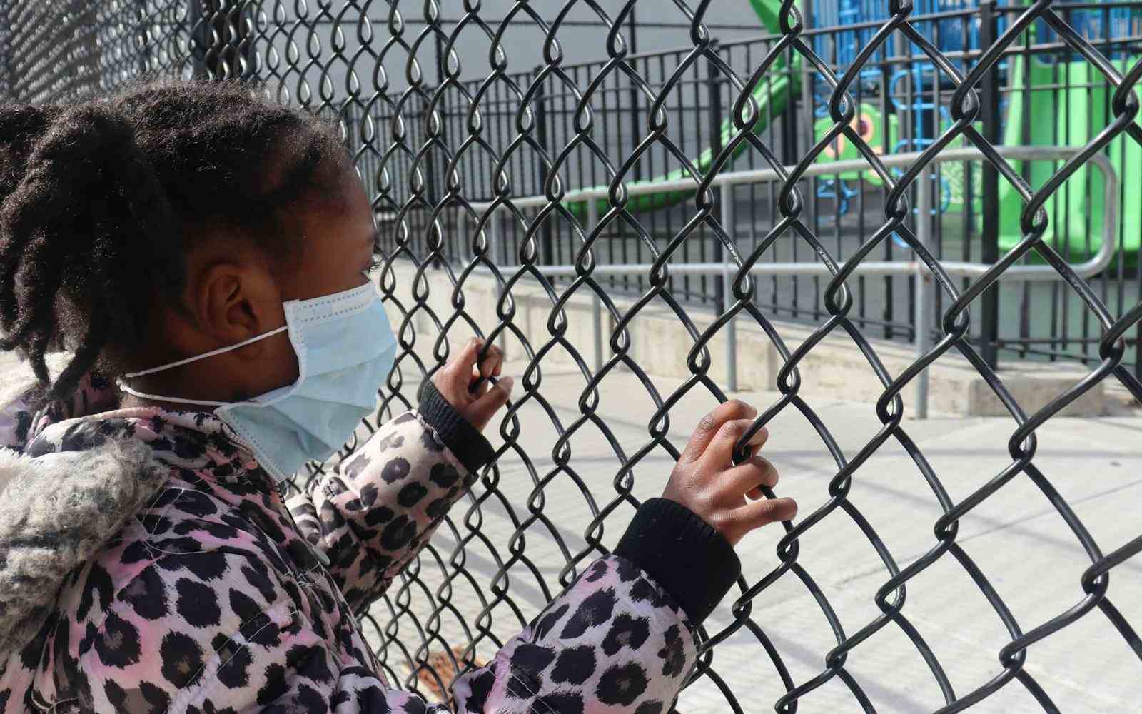 young child with hands on a chain-link fence looking at a playground inside the fence