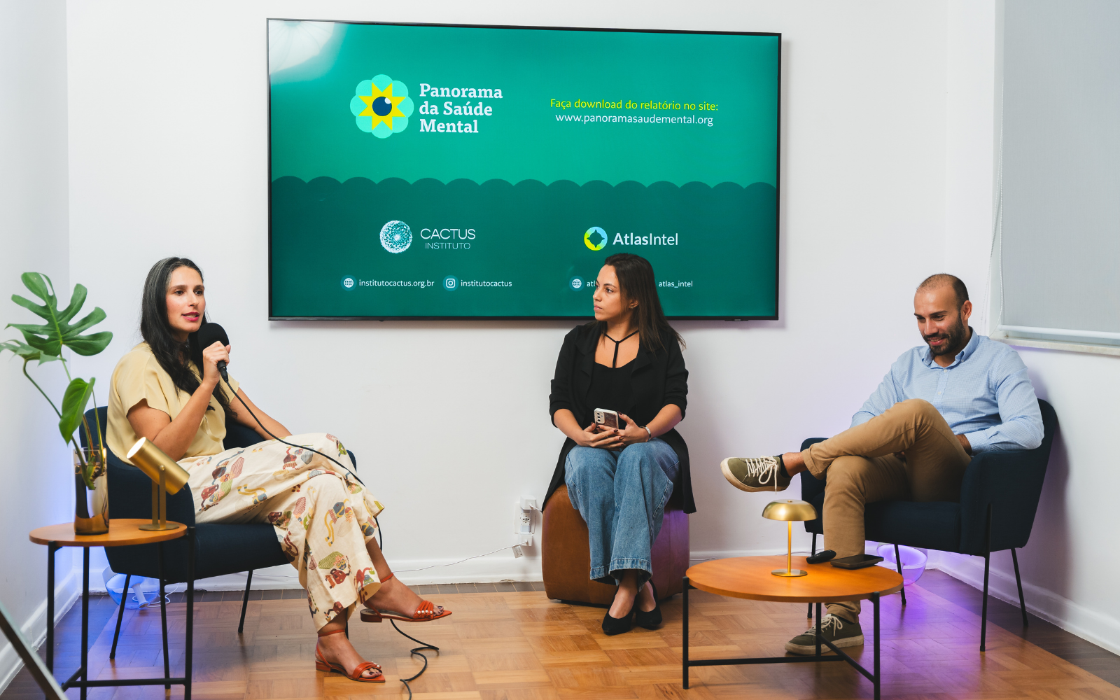 Três pessoas sentadas em poltronas falam em um painel. Atrás delas, há uma televisão que dá destaque ao Panorama da Saúde Mental