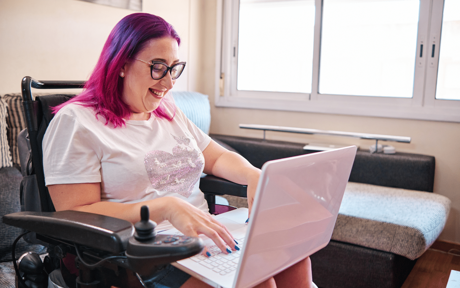A person with pink hair uses a laptop while seated in a powered wheelchair in a bright living room.