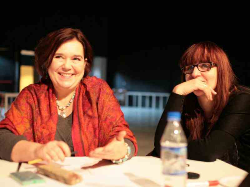 Zdjęcie przedstawia dwie uśmiechnięte kobiety siedzące przy stole. Po lewej Katarzyna Oleś w czerwonej chuście na ramionach i kolorowym naszyjniku.