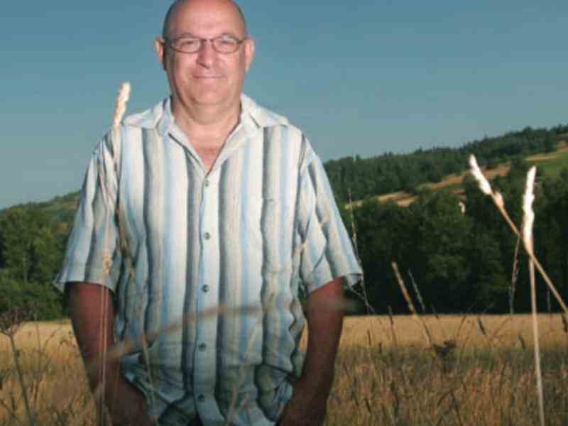 Zdjęcia Piotra Topińskiego na tle pola. Piotr ubrany jest w białą koszulę w pionowe pasy. Uśmiecha się.