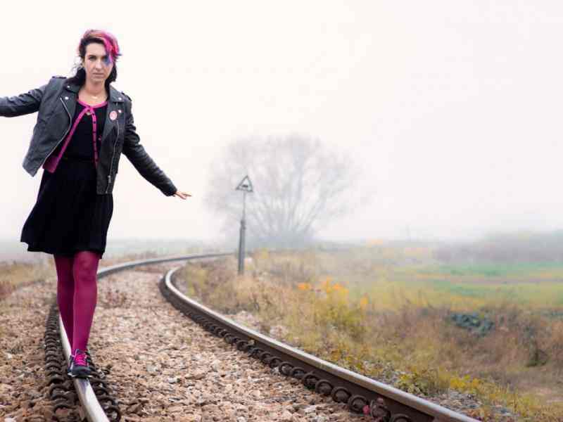 Zdjęcie Sylwii Chutnik idącej po torach kolejowych biegnących przez pole. W tle mgła. Sylwia ma różowe pasemko we włosach i różowe rajstopy.