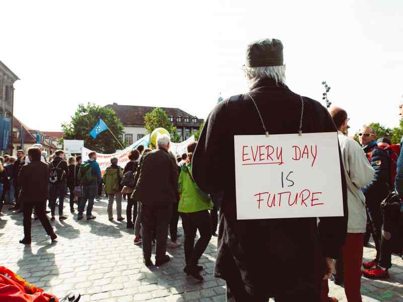 Mężczyzna w ciemnej kurtce ma na plecach zawieszony baner z napisem "every day is future". W tle widać innych protestujących,