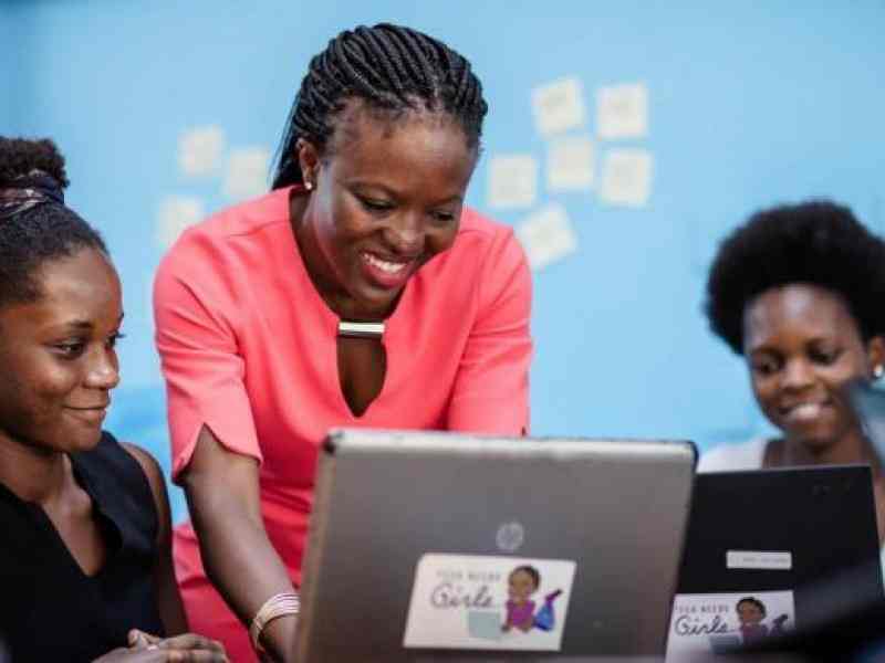 Na zdjęciu 3 kobiety wspólnie pracują przy laptopach. Uśmiechają się za nimi jasno-niebieska ściana.