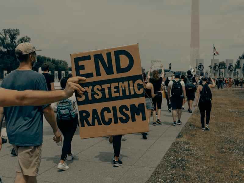 Karton z napisem "end systemic racism" (tłum. zakończmy systemowy rasizm) trzymana przez osobę protestującą. W tle inne osoby uczestniczące w proteście.