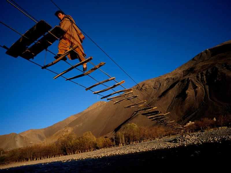 2020 Leading Social Entrepreneurs Front Cover. Depicting a man in the valleys of northeastern Afghanistan crossing a cable bridge with sparsely spaced wood planks above the Pandjchir River, connecting the village of Malespe to a refugee camp. Photographer and Ashoka Fellow, Reza Deghati, captured this shot in 2000.