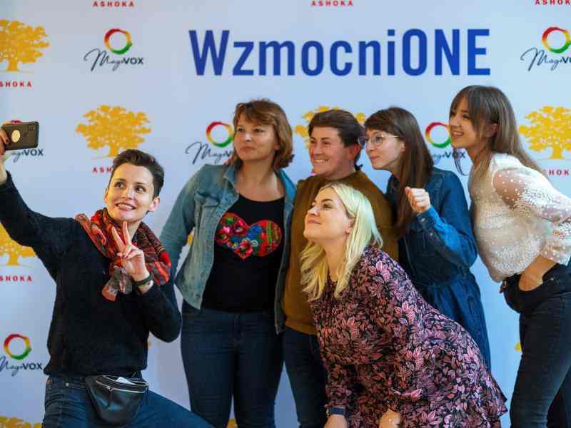 Na tle ścianki zdjęciowej z napisem Wzmocnione oraz logo Ashoki i Magovox 6 kobiet robi sobie selfie.
